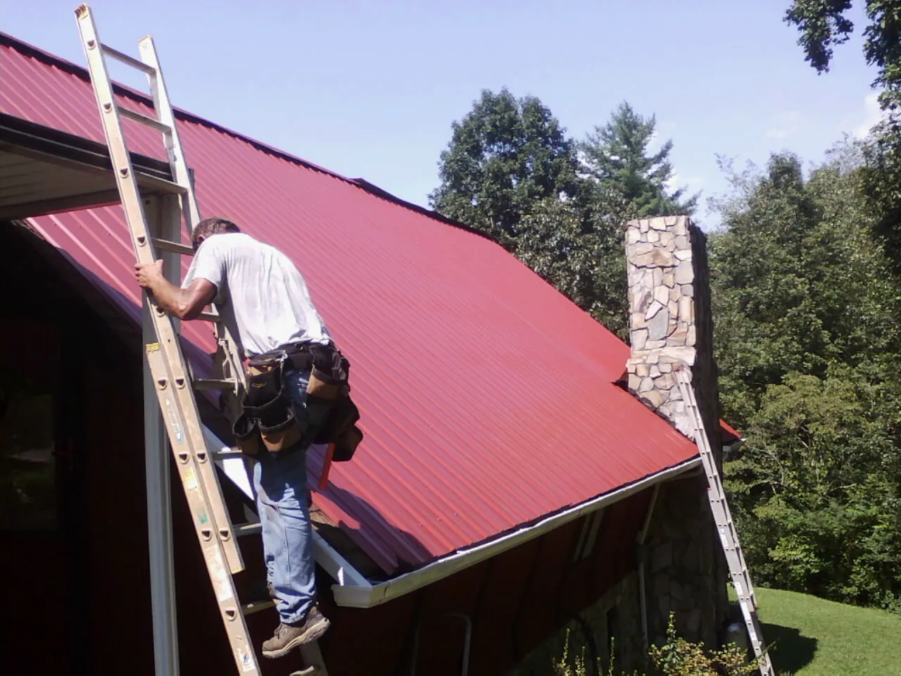 Roofer on a ladder beside a red metal roof and stone chimney — Soffit & Fascia Repair by Roofing Brothers in Eastlake